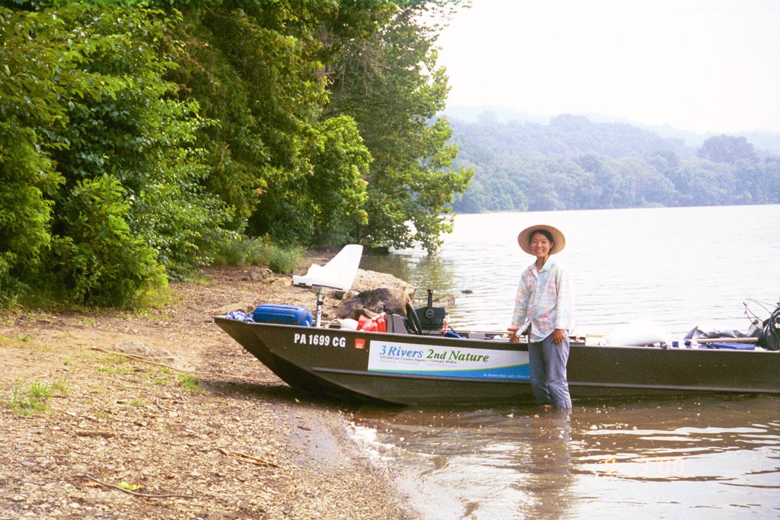 Reiko Goto standing in the shallows beside the 3 Rivers 2nd Nature survey boat, pulled up on a wooded riverbank